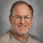 Man with glasses, light beard, and short hair, wearing a gray shirt, smiling against a gray background.