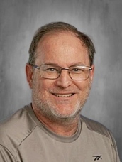 Man with glasses, light beard, and short hair, wearing a gray shirt, smiling against a gray background.