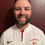 Smiling man with a beard wearing a white sports jacket with a logo, standing against a maroon wall.