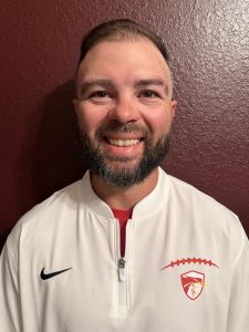 Smiling man with a beard wearing a white sports jacket with a logo, standing against a maroon wall.