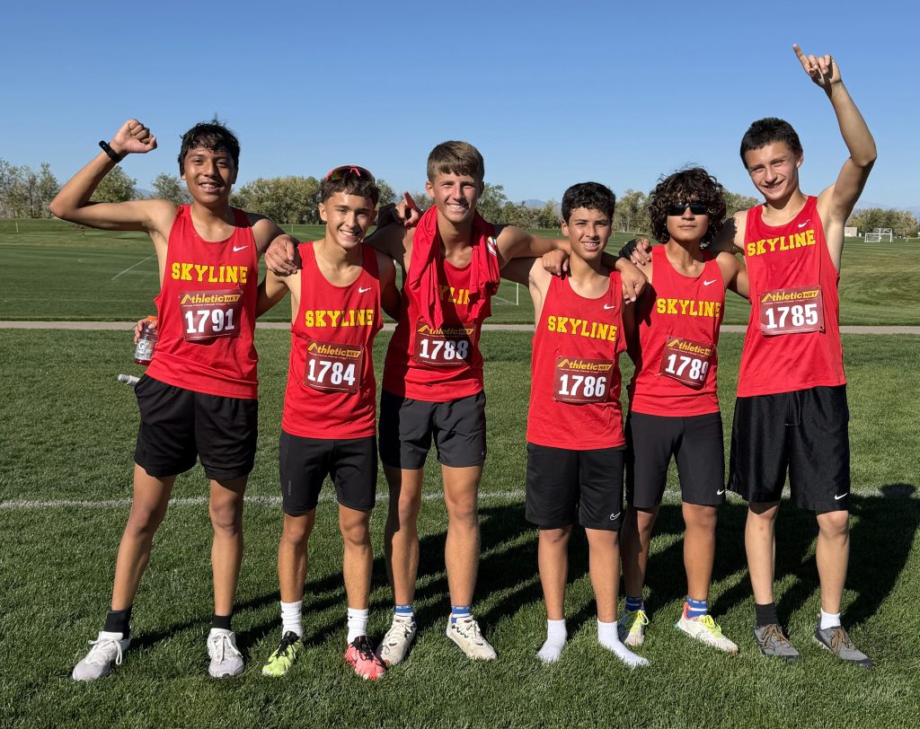 Six cross-country runners in red jerseys pose on a grassy field, smiling with arms around each other.