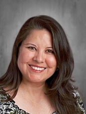 Smiling woman with long dark hair, wearing a patterned blouse, against a gray background.
